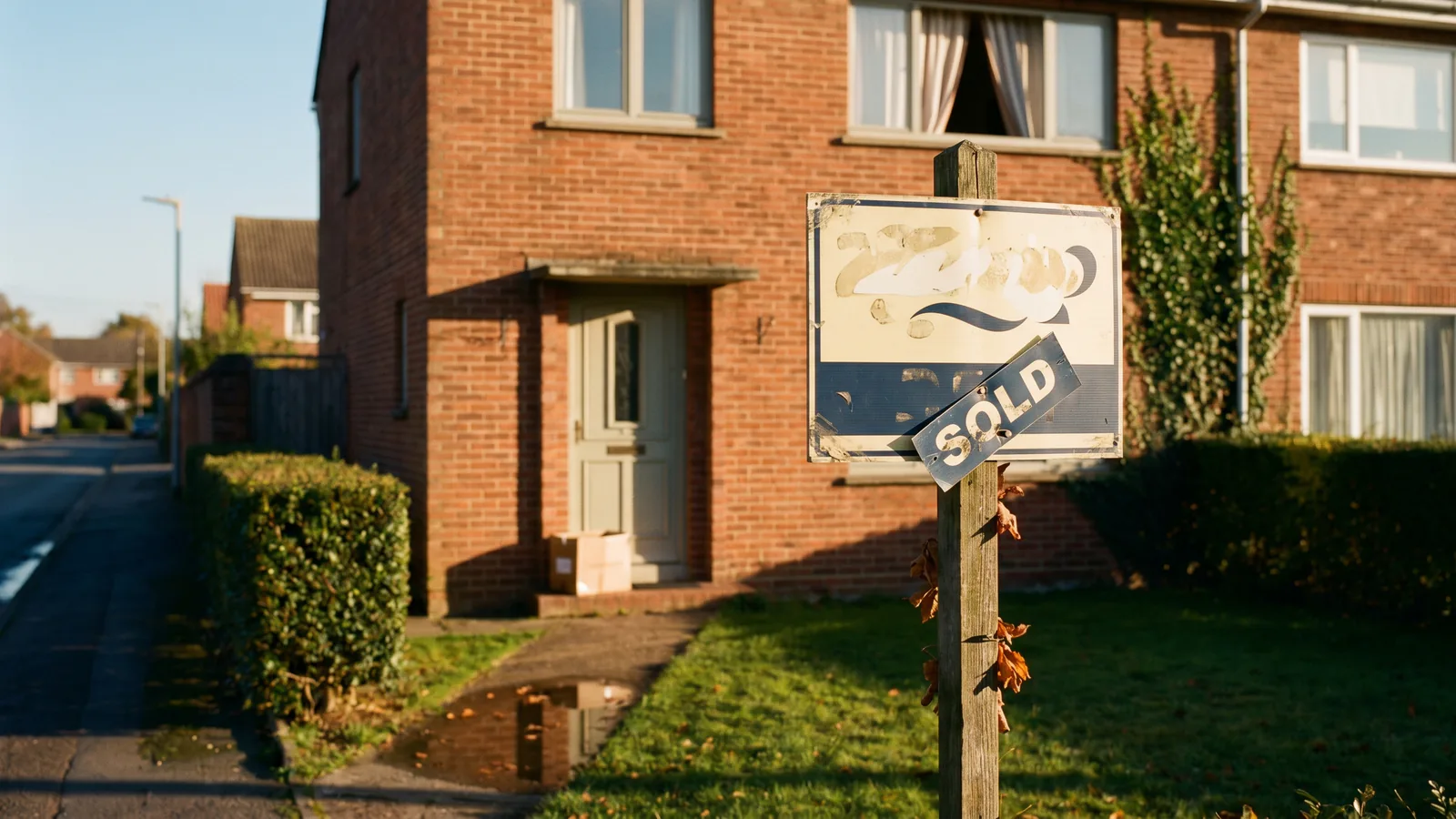 The front of a modest European semi-detached brick home in late golden-hour light with a weathered wooden estate-agent For Sale board planted in the small front garden, ivy on one corner of the brickwork, autumn leaves around the sign post, no people.