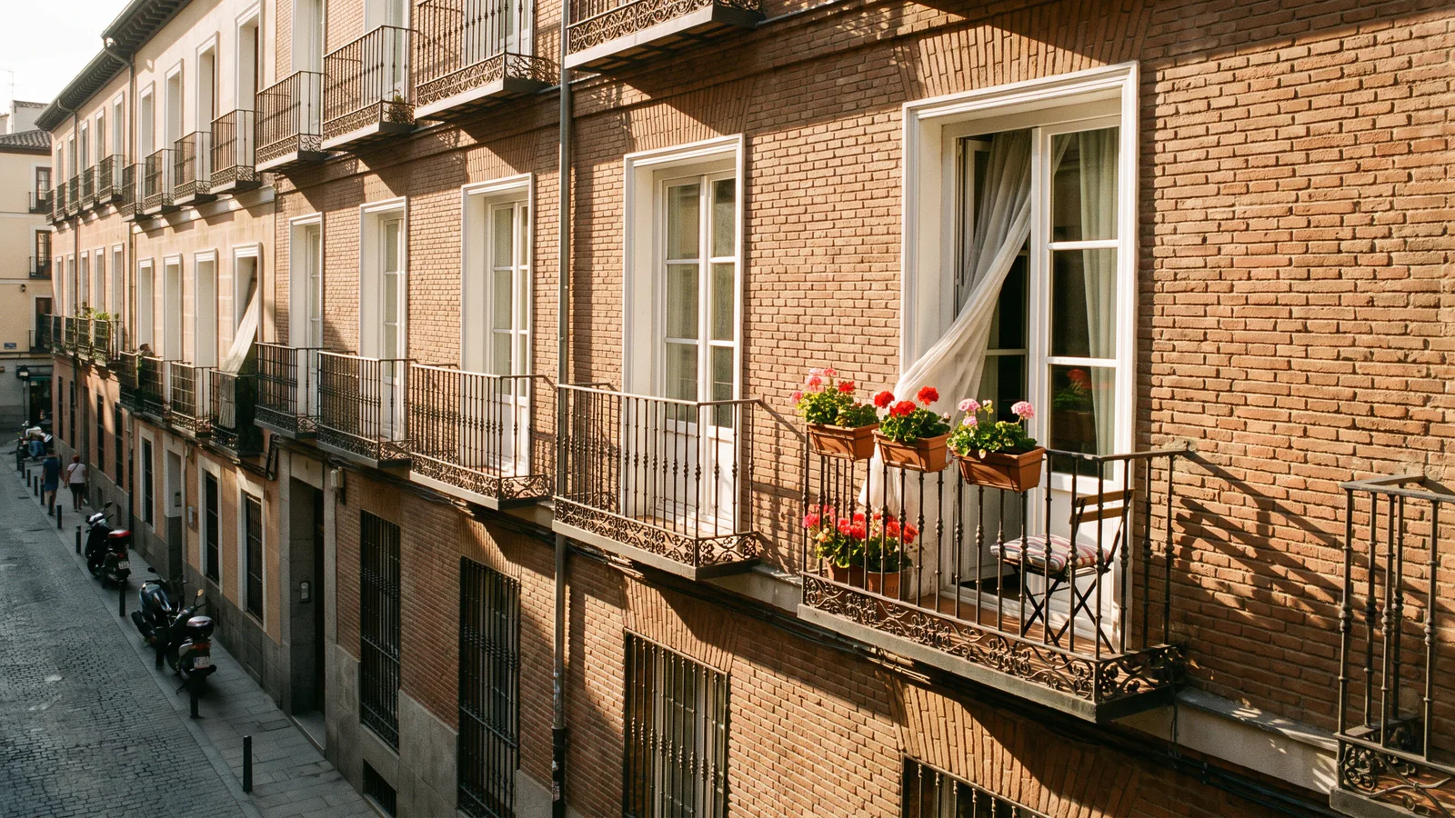 Warm brick facade of a classic Madrid apartment building with white-framed windows and wrought-iron balconies in late-afternoon sunlight, one balcony holding potted geraniums and a small folding chair.