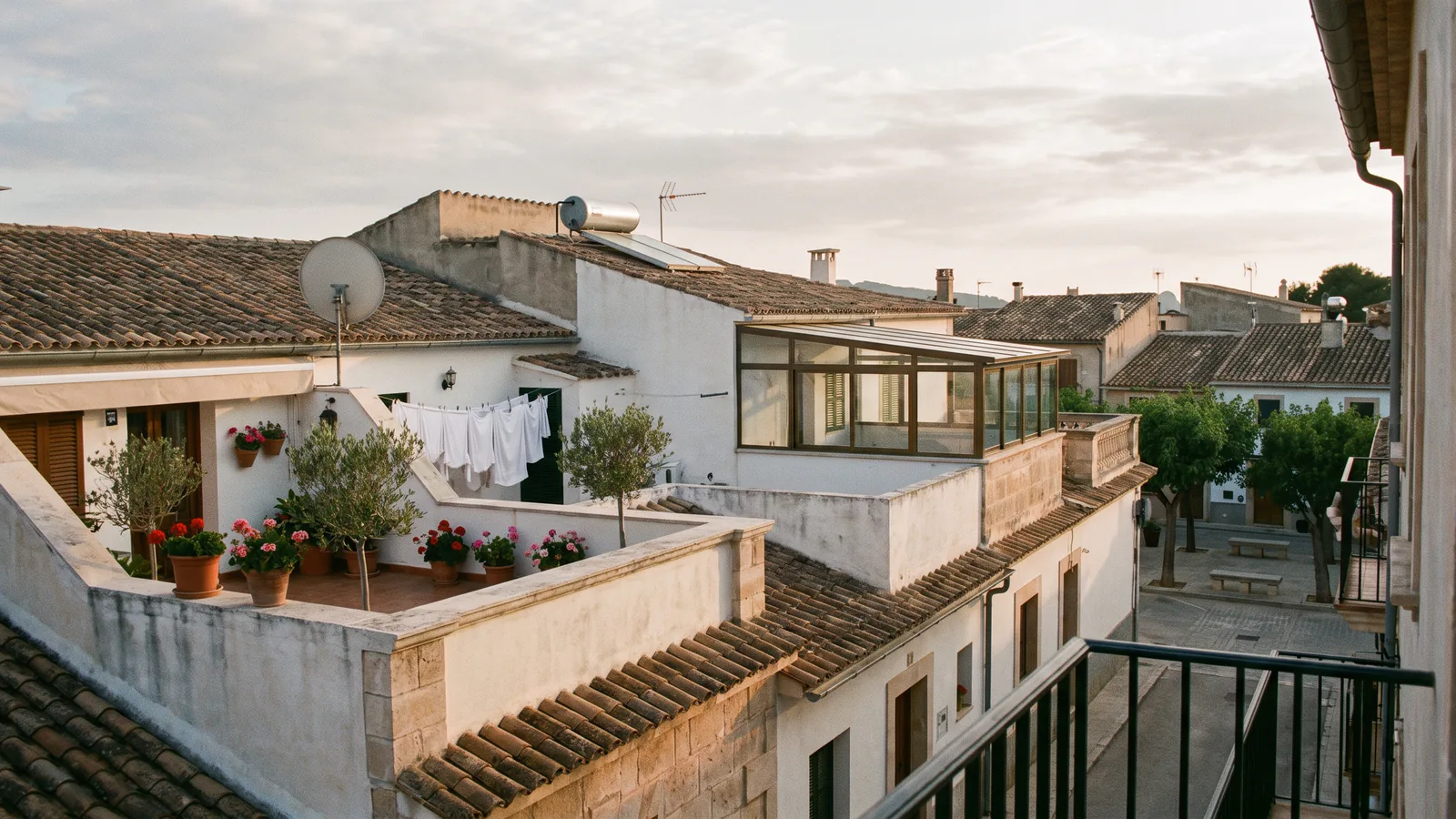 Vista elevada sobre los tejados y las terrazas privadas de un edificio mediterráneo bien conservado, con teja árabe, plantas en macetas y una galería acristalada añadida por encima de la línea original del tejado, bajo la luz suave del atardecer.