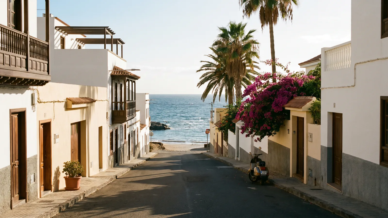 Uma rua residencial costeira e soalheira de Tenerife que desce suavemente para uma praia de areia e o Atlântico azul, ladeada por casas canárias bem conservadas em branco e creme, com varandas de madeira escura e palmeiras.