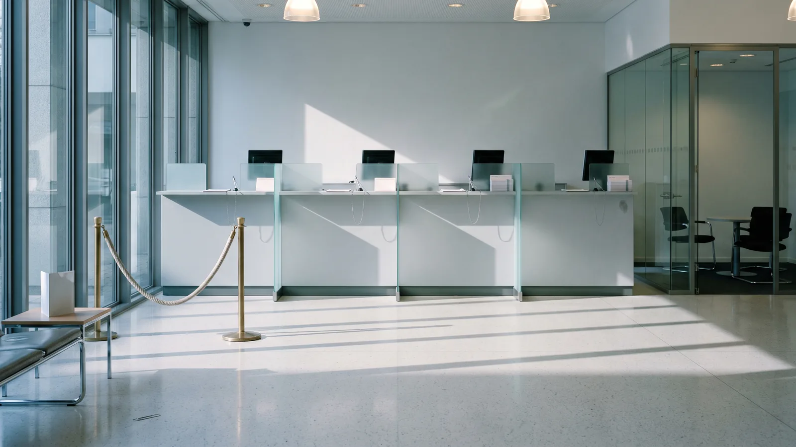 The interior of a modern European bank branch early in the morning before opening — a row of teller counter stations with frosted-glass dividers, a glass-walled consultation office visible in the background, a low waiting bench and queue stanchion in the foreground, and cool morning daylight through tall windows.